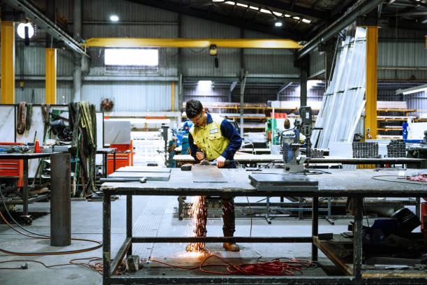 istock Industrial worker in a workshop wearing safety gear using a grinder emitting sparkse in large industrial warehouse with machinery and yellow support structures under bright overhead lighting 2188581309