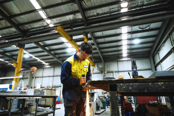 istock Industrial worker in a workshop wearing safety gear using a grinder emitting sparkse in large industrial warehouse with machinery and yellow support structures under bright overhead lighting 2188581310