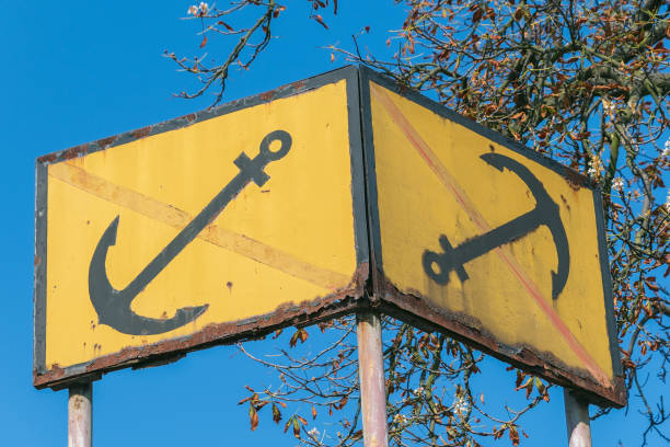 Rusty yellow no-anchoring sign featuring black anchors, against a blue sky and tree branches.