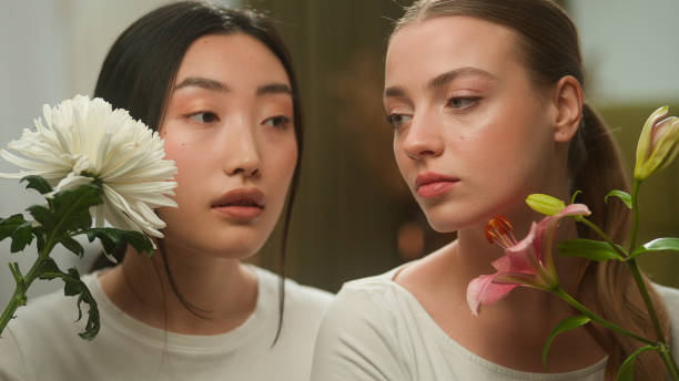 Two beautiful healthy multiracial girls floral bloom organic cosmetic aroma perfume cosmetology make-up multiethnic females women Asian woman girl Caucasian holding flowers near faces skin care beauty. High quality 4k footage dos hermosas chicas multirraciales saludables floral flor cosmética orgánica aroma perfume cosmetología maquillaje mujeres multiétnicas mujeres mujer asiática niña caucásica sosteniendo flores cerca de las caras cuidado de la piel belleza