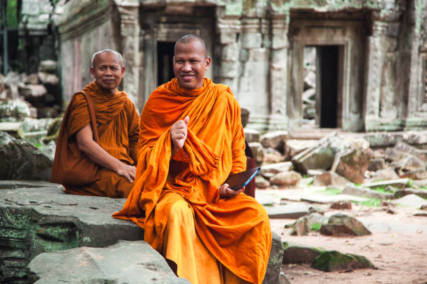 Two monks in vibrant orange robes sit among the stone ruins of Cambodia, enjoying a moment of conversation. The peaceful atmosphere highlights the cultural richness of the area.