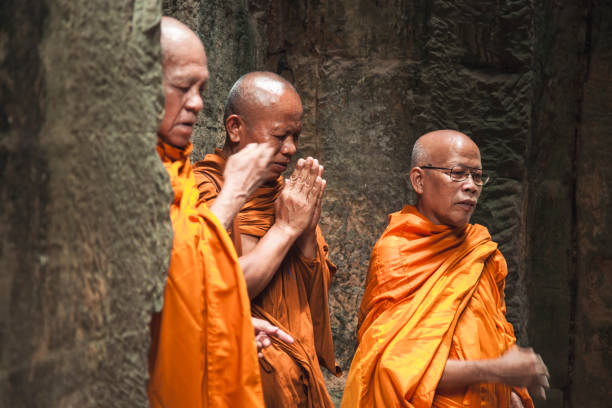 Angkor Wat, Cambodia - April 19, 2022: A young monk in an orange robe walks along the stone pathway of Angkor Wat as tourists meander through the historic site in the morning light, enjoying the scenery.