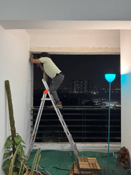 Stock photo showing close-up, profile view of an Indian handyman, wearing work boots, standing on the rung of an aluminium step ladder on a high apartment balcony, whilst he leans precariously to install anti-bird netting. Health and safety concept. imagen de un manitas irreconocible instalando una red anti-pájaros para palomas en un balcón residencial alto, apoyado precariamente en una escalera de mano, fondo de expansión urbana, noche, vista de perfil, enfoque en primer plano, concepto de salud y
