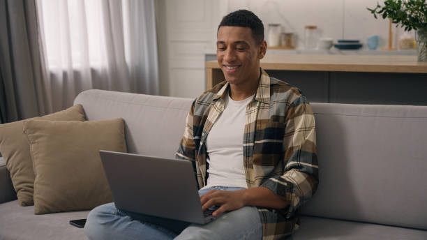 istock Happy smiling African American ethnic man working at home distant online work studying at sofa guy laptop using computer at couch male using pc playing internet video game use app shopping chatting 2188650972