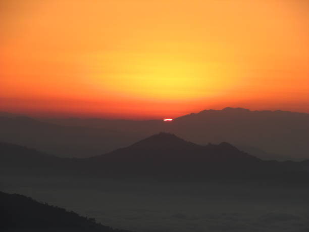 The picture is a landscape image.It depicts a sunrise over a mountain range.The sky is a vibrant orange and yellow, and the sun is just beginning to peek over the horizon.The mountains are silhouetted against the sky, and there is a layer of fog in the foreground.The image conveys a sense of peace and tranquility.