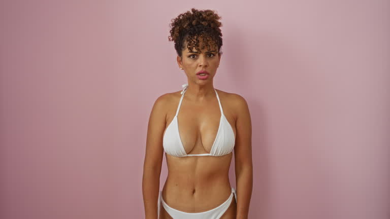 Young hispanic woman in bikini standing and looking skeptical with sarcastic open-mouth expression over isolated pink background