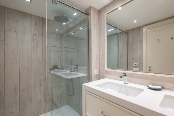 istock Spacious bathroom with bright artificial lighting, large mirror reflecting front door and pair of snow-white sinks built into light beige cabinet. Shower enclosure with blue glass splash guard. 2188678633