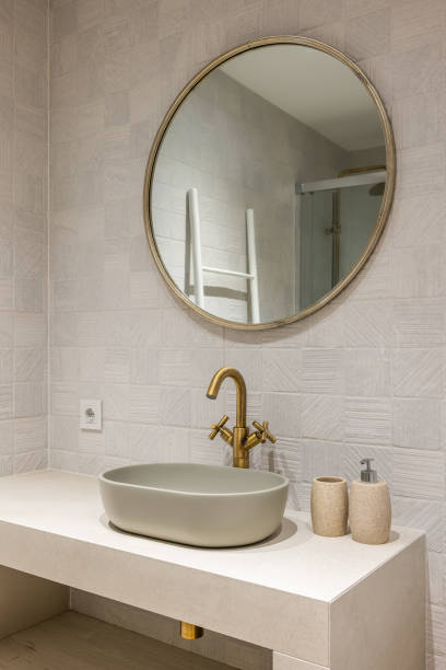 istock Neat stylish oval-shaped sink on white marble countertop with an elegant copper-coloured faucet. Above sink hangs a round modern gold-framed mirror that reflects part of shower. 2188678903