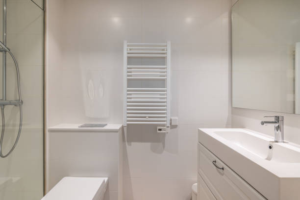 Bathroom in light colors with glossy ceramic tile walls and bright lighting. The bathroom has a shower with a glass railing, a large mirror with a sink, a radiator for drying towels