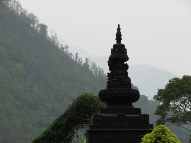 The picture showcases a black stone monument, resembling a stupa or a spire, situated against a backdrop of lush green hills.\nThe monument is adorned with intricate carvings and figures.\nThe silhouette of the monument stands out against the misty and hazy background, creating a sense of mystery and intrigue.\nThe overall tone of the picture is serene and tranquil, evoking a sense of peace and contemplation.
