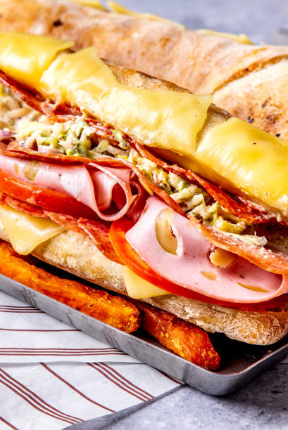Viral Grinder sandwich with deli meat - pepperonis, ham, mortadella, grilled sliced cheese, stuffing with cabbage served in a French baguette with sweet potato fries as a side dish on a metal pan