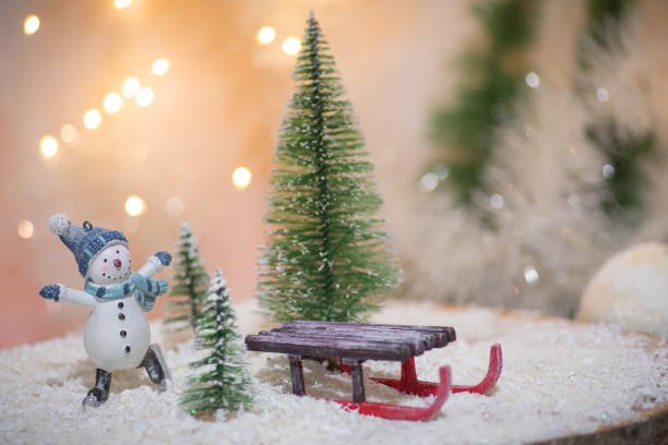 A charming winter wonderland is depicted with a tiny snowman dressed warmly, accompanied by a small sled surrounded by miniature pine trees and fake snow.Christmas background with copy space