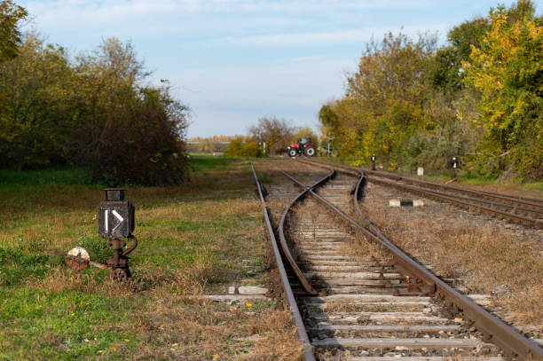 A rural railroad scene in autumn, showing tracks with a point indicator in the foreground. A tractor and hay bales are visible in the distance, blending agricultural and railway elements in a tranquil countryside setting.