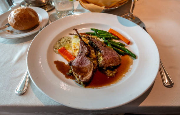 Gourmet dish featuring herb-crusted lamb chops with gravy, roasted vegetables, green beans, mashed potatoes, and creamy sauce, served on a white plate with bread and a drink on a formal table setting
