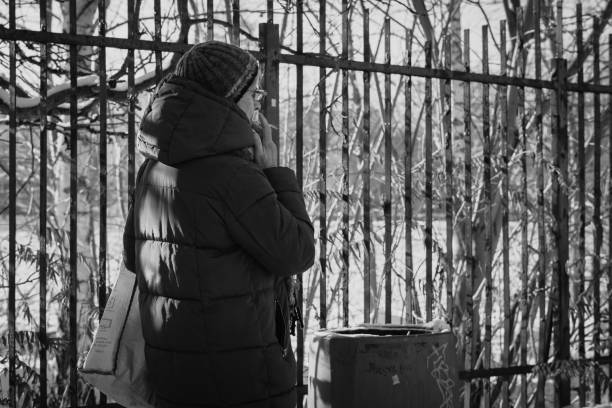 Krakow, Poland - November 30th 2023: A woman in a thick winter coat and hat stands near a snow-covered iron fence, smoking quietly on a cold day in Krakow, Poland. The sunlight filters through bare branches behind her, creating a tranquil and reflective mood. The combination of nature, urban elements, and solitude captures a moment of calm during the winter season.