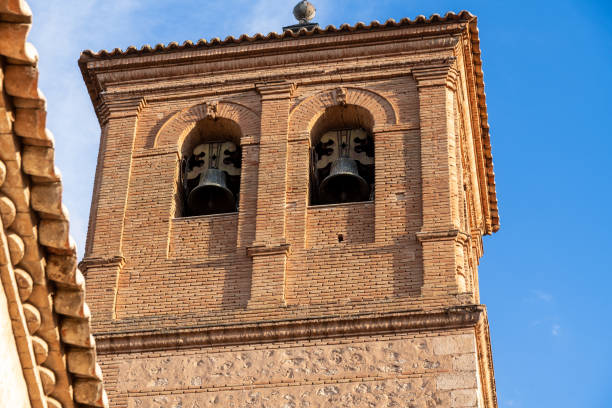 Red brick bell tower with two arches, bells, decorative elements, and a clear blue sky with part of a roof visible.