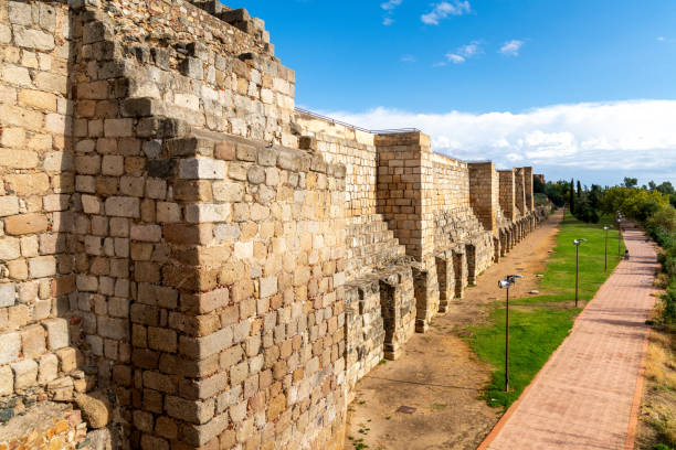 Massive stone walls of the Alcazaba in Merida with regular battlements, surrounded by a path and greenery. Sky with clouds and a blue background.