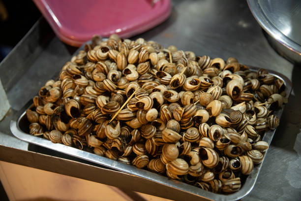 A metal bowl filled with cooked snails, featuring brown and cream striped patterns, with blurry kitchen utensils in the background.