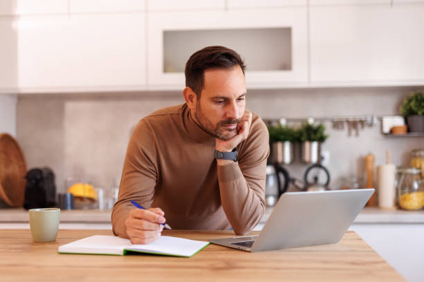 istock Serious man with hand on chin reading data over laptop while writing in note pad on table in kitchen 2188753880