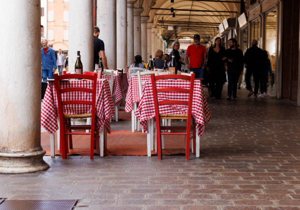 Mantova, Italy - September 15, 2024: Empty tables and chairs outside a restaurant under the city's iconic porticos, with tables featuring the classic Italian red and white checkered tablecloths