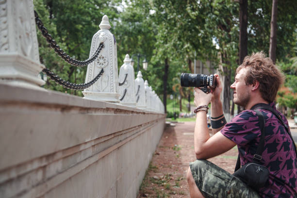 In Phnom Penh, a photographer kneels beside a decorative stone fence, focusing intently with a DSLR camera, surrounded by lush greenery and a tranquil atmosphere during a sunny day.