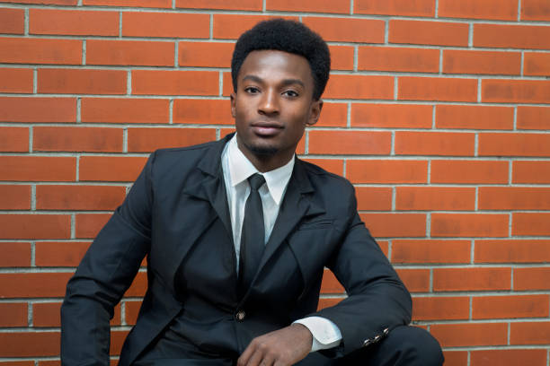 Portrait of a confident young man in a black suit and tie, sitting against a brick wall, showcasing professionalism, style, and a modern, polished look.