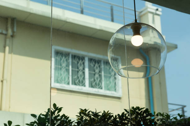 A lamp with a bulb in a glass globe hangs over a window. The lamp is lit up, and the window is open. The scene is bright and cheerful, with the sunlight streaming in through the window