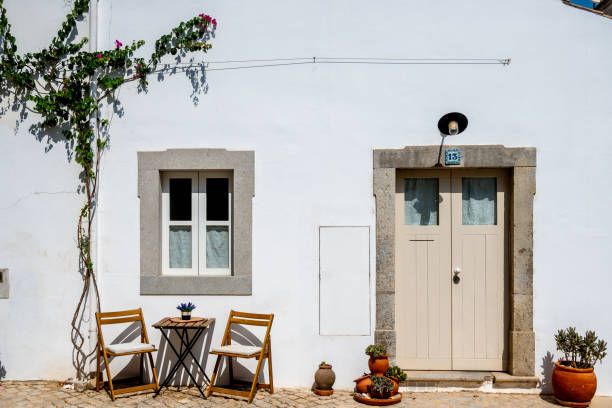 Tavira, Algarve - Portugal - 09-18-2024: White house facade with a door and windows, flanked by potted plants and a small table with two chairs on a paved floor.
