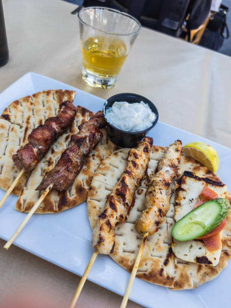 A delicious serving of traditional Cypriot souvlaki on pita bread, accompanied by grilled halloumi, tzatziki, and fresh vegetables, paired with a glass of local drink.