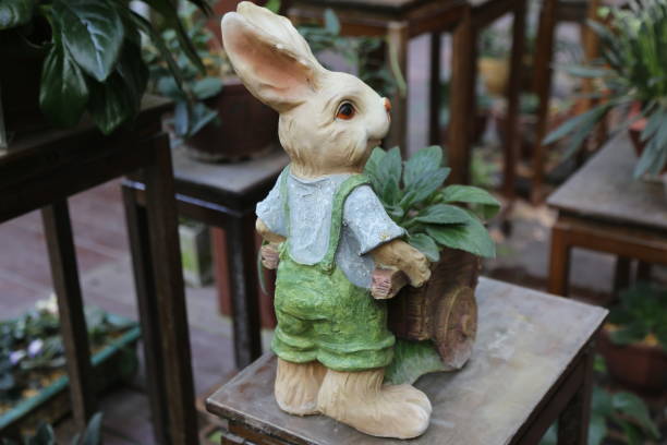 A rabbit is holding a plant in a pot. The rabbit is wearing a green apron and holding a small shovel. The scene is set in a greenhouse with many potted plants