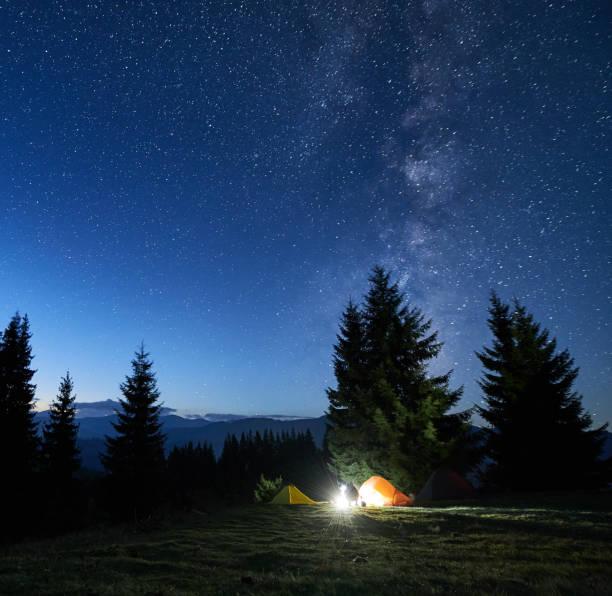 istock Amidst forested mountain landscape, two tents glow warmly beside campfire under star-filled sky. 2188869513