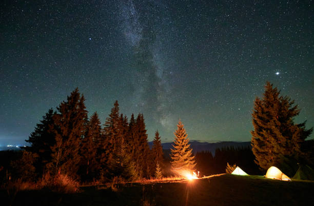 istock Amidst forested mountain landscape, two tents glow warmly beside campfire under star-filled sky. 2188869514