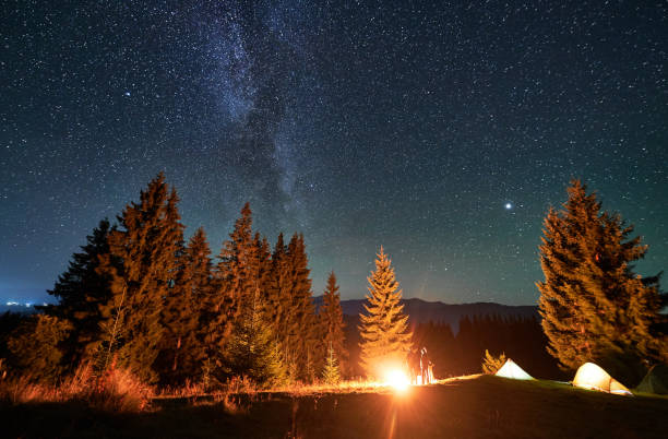 istock Amidst forested mountain landscape, two tents glow warmly beside campfire under star-filled sky. 2188869523