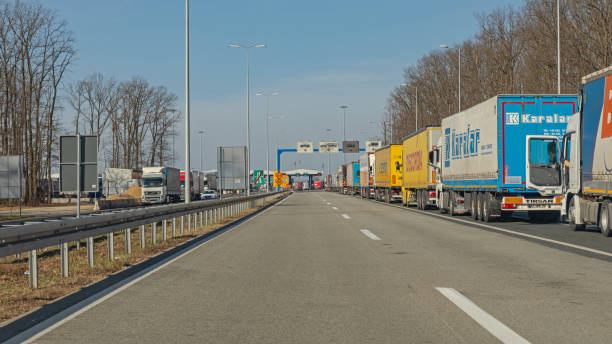 A long line of cargo trucks waits at a highway checkpoint under a clear sky, with trees and road signs in the background.
