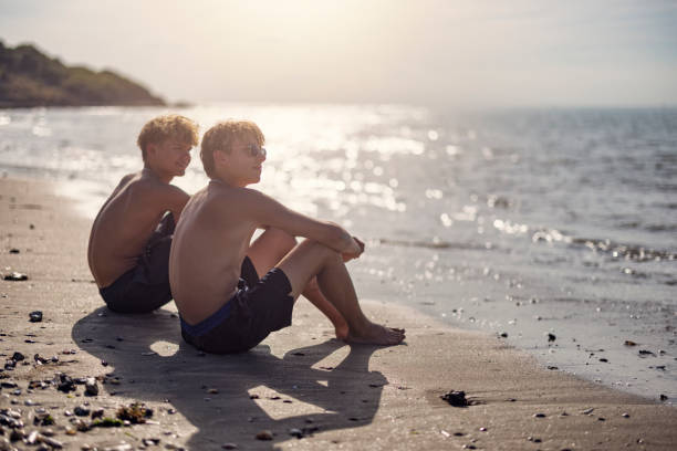 Two teenagers are sitting on a wide sandy beach in Calvados, Normandy, France. The sand is scattered with seashells.\nShot with Canon R5, on a sunny summer day.