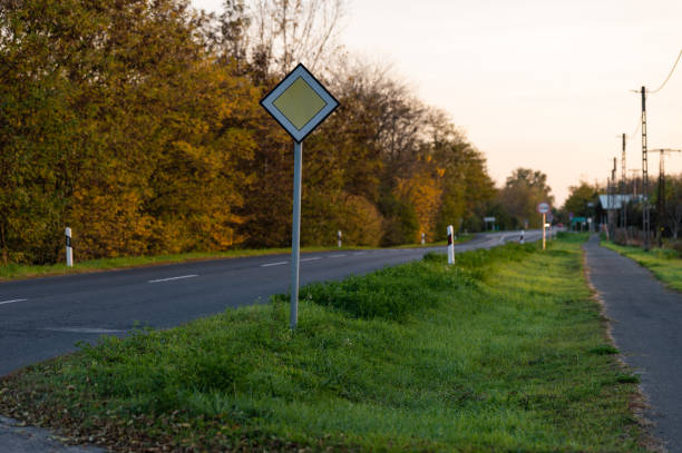 A diamond-shaped priority road sign along a rural road lined with autumn foliage. The scene captures the quiet ambiance of the countryside with lush greenery and golden leaves in the background.