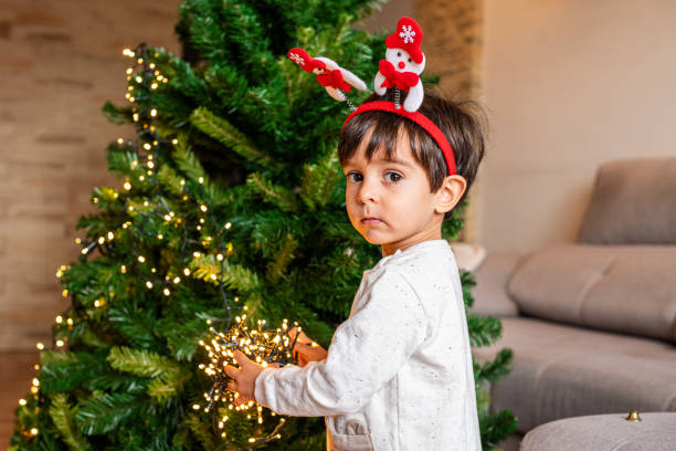 istock A young boy is standing in front of a Christmas tree, wearing a Santa hat and holding a string of lights 2188890045