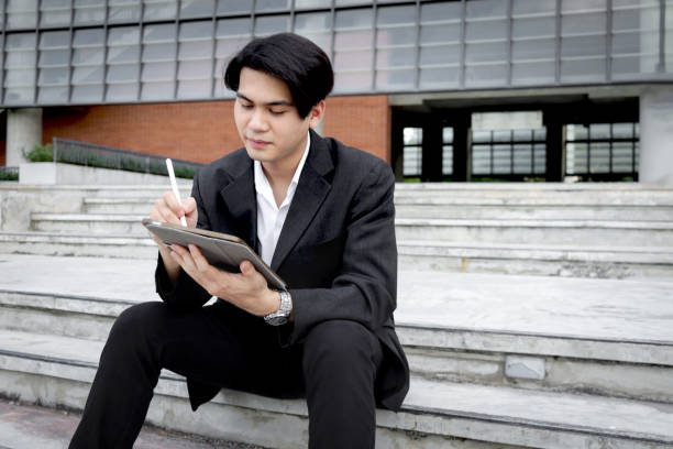 istock Asian young businessman in black suit using digital tablet for working while sits on a railing in front of modern building. Male officer using tablet outdoor office. Happy smart businessman using electronic devices outdoors in urban modern environments. 2188895228
