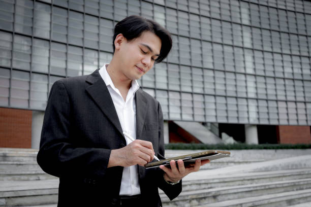 istock Asian young businessman in black suit using digital tablet for working while stands in front of modern building. Male officer using tablet outdoor office. Happy smart businessman using electronic devices outdoors in urban modern environments. 2188895470