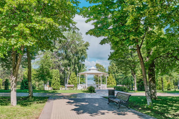 A picturesque white gazebo in a lush green Park - Strukovsky Garden, Samara, Russia. Paved walkways and benches, framed by tall trees. View of the Volga River. Summer landscape. Peaceful setting
