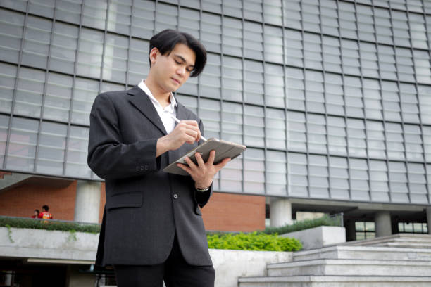istock Asian young businessman in black suit using digital tablet for working while standing in front of modern building. Male officer using tablet outdoor office. Happy smart businessman using electronic devices outdoors in urban modern environments. 2188898847