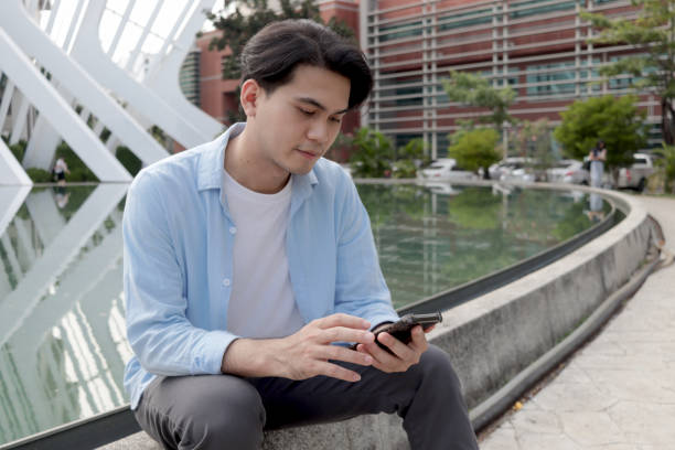 istock Asian young man casual clothes looks at mobile phone and texts message while siting in front of modern building. Male using smartphone in outdoor. Happy smart young man using electronic devices outdoors in urban modern environments. 2188900031