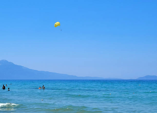 Aydın, Turkey - July 18, 2024: People enjoying near the sea at beach on summer holiday. Some of them swimming and others are parasailing on Aegean sea, you see town of Kuşadası in Turkey on a sunny summer day, View Of Kusadasi Skyline At Aegean Coast, Turkey.