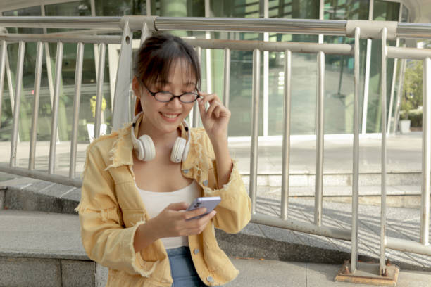 istock Asian young woman looks at her mobile phone and texting message while sits on a railing in front of modern building. Female uses smartphone in city. Happy woman using electronic devices outdoors in urban modern environments. 2188908295