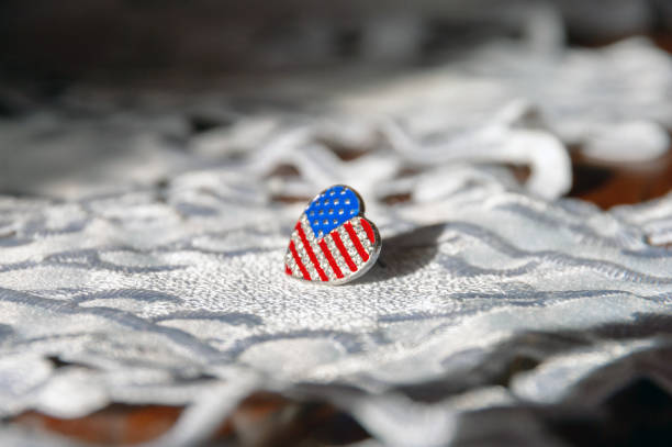 A heart shaped American flag pin sits on a white cloth. The flag pin is made of silver and has a patriotic design. Concept of patriotism and pride in one's country