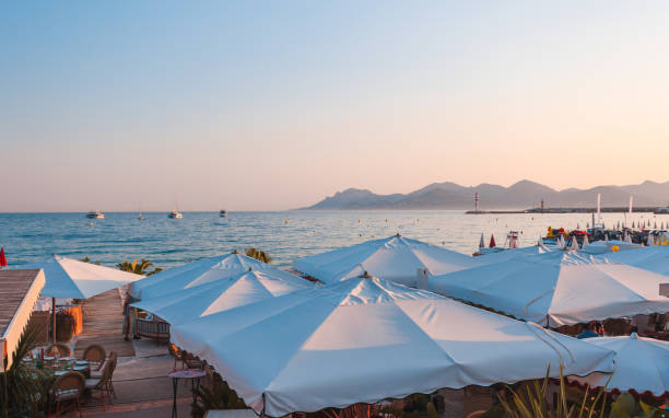 Seaside view at sunset in Cannes, French Riviera, with white parasols, a calm sea with yachts, mountains, and a lighthouse on the horizon. The warm candid hues of the sky and tranquil ambiance capture the essence of a Mediterranean summer evening.