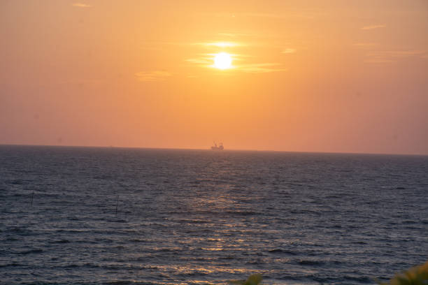 A mesmerizing sunset paints the sky and sea in golden hues, evoking a sense of tranquility. In the distance, a lone ship floats on the calm waters, silhouetted against the radiant glow of the setting sun. The minimalistic composition emphasizes the vastness of the ocean and the serene beauty of nature at day's end.