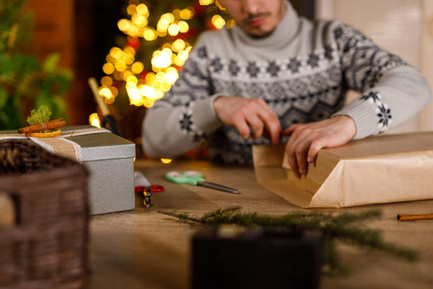 istock Young man folding edges of a rustic brown paper that he is using to wrap a Christmas gift box 2188932333