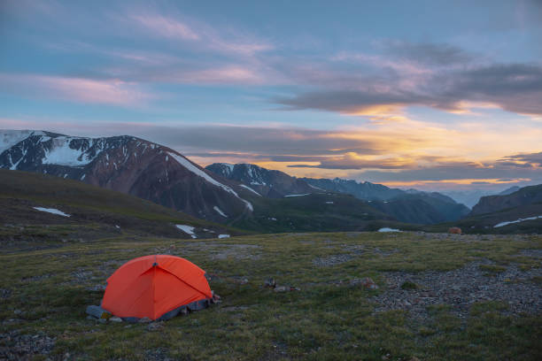 istock Orange tent on grassy stony pass with view to mountain range silhouette far away under clouds in vivid sunset color sky. Scenic alpine landscape with red tent in mountains under colorful cloudy sky. 2188932359