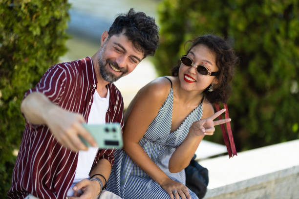 istock Happy couple taking a cheerful selfie together outdoors with trees in the background, both smiling and making peace signs while enjoying a sunny day. 2188935513